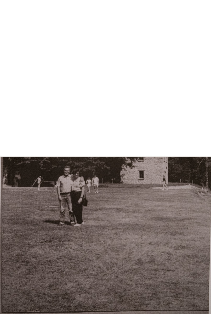 Foto vintage en blanco y negro de una pareja sonriente de pie al aire libre, con árboles y personas al fondo.