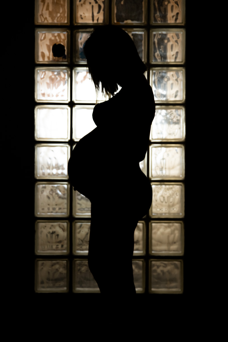 Silhouette of a pregnant woman standing sideways in front of a window with glass blocks.