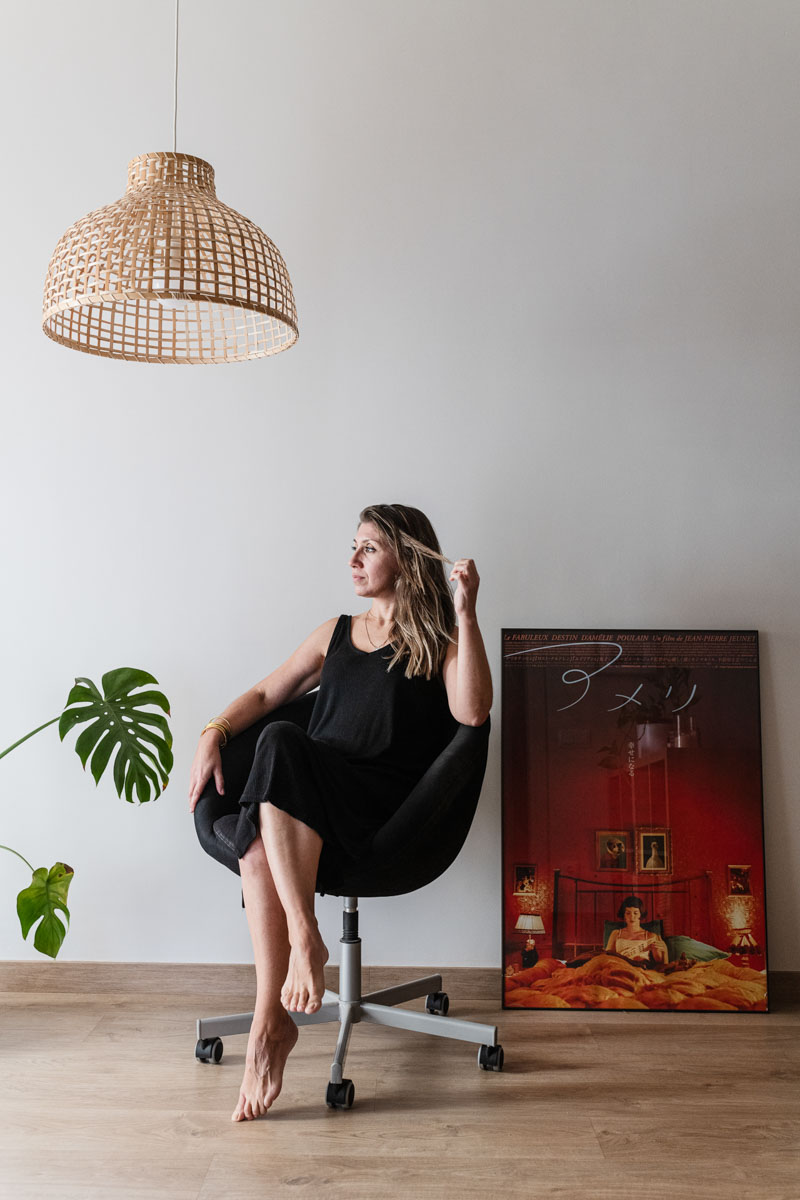 Woman in a black dress sitting barefoot on a black office chair, with a wooden floor, a large green plant, a woven pendant light, and a framed artwork leaning against the wall.