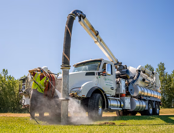 Worker with hose operating white vacuum truck on green grass with trees and clear sky background.