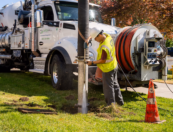 Worker in safety vest uses vacuum truck hose for outdoor maintenance near open ground and traffic cone.
