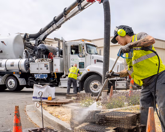 Worker in safety gear pressure washing metal grates near vacuum truck at construction site.