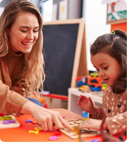 Smiling woman helps young girl with a wooden puzzle at a colorful table in a classroom.