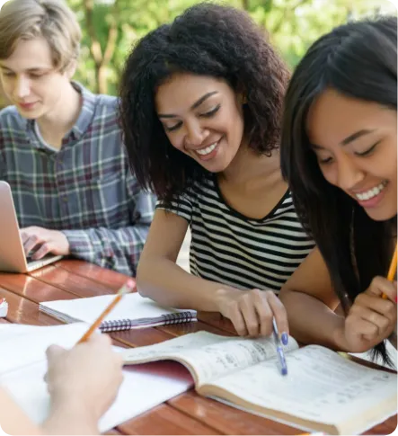 Three students studying together outdoors, looking at open books and notes on a wooden table.