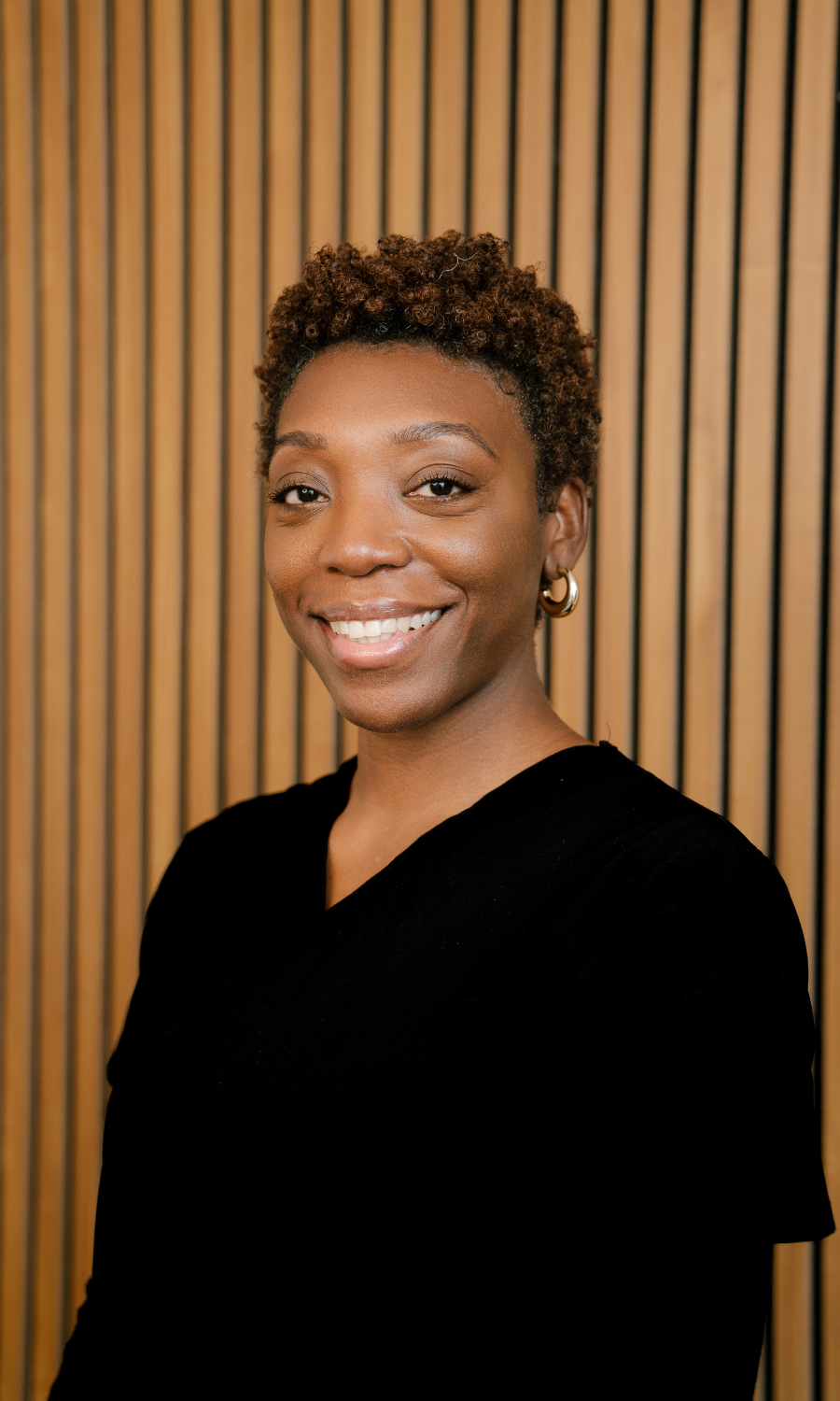 Smiling woman with short curly hair wearing gold hoop earrings and a black top in front of a wooden slat background.