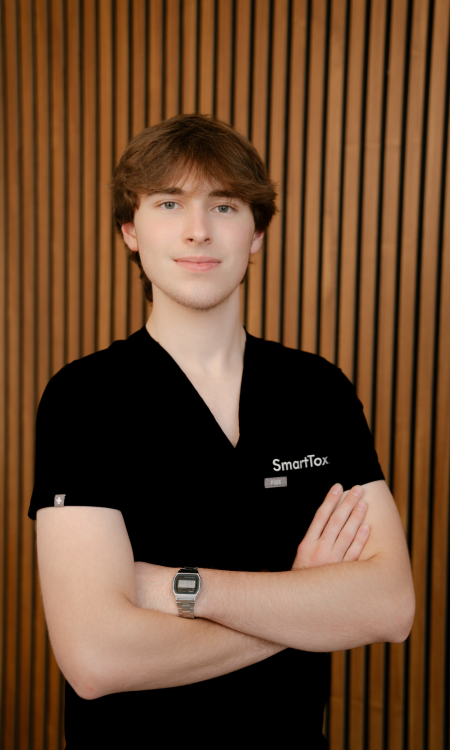 Young man with light brown hair wearing a black SmartTox shirt and a digital wristwatch, standing with arms crossed in front of a vertical wooden slat wall.