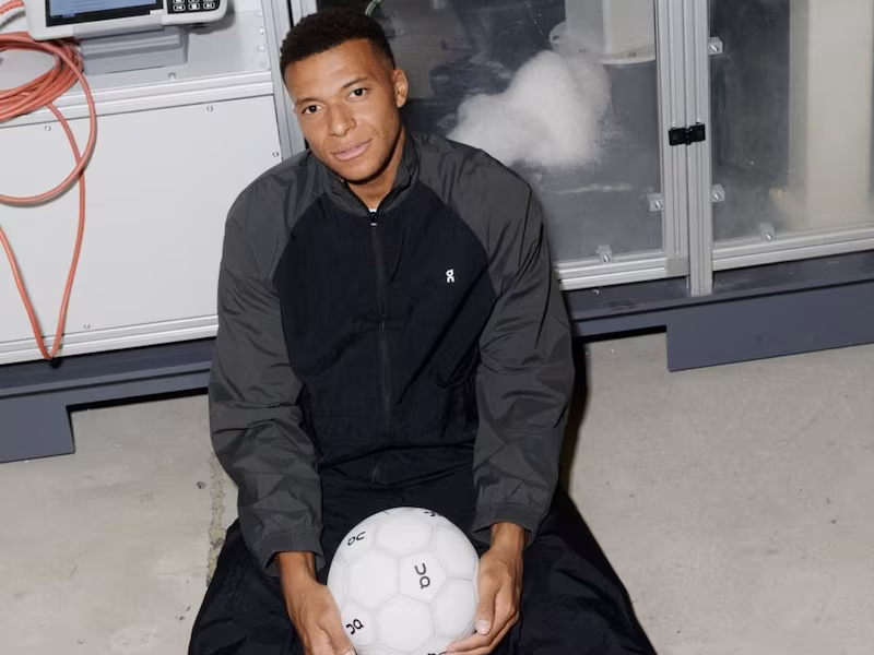 Mbappe sentado en el piso de un laboratorio sostiene una pelota de fútbol blanca con el logo de On y viste una campera gris y negra de la marca suiza.