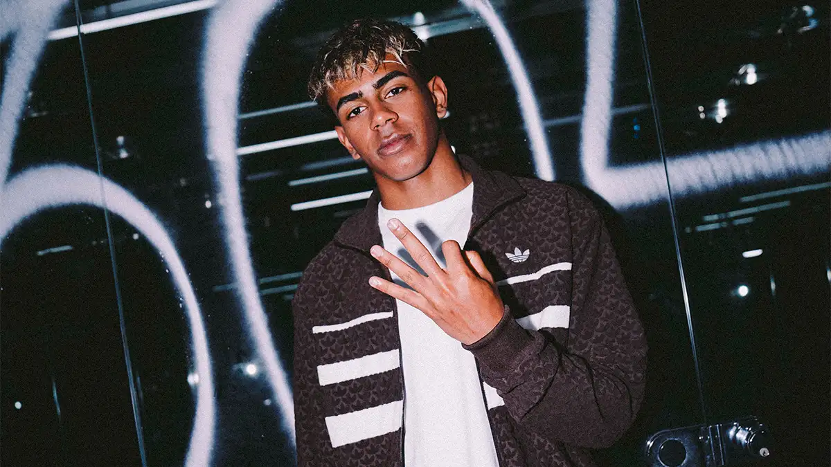 Lamine Yamal posa con una campera marrón de Adidas con rayas blancas y una remera blanca, frente a un fondo oscuro con grafitis.