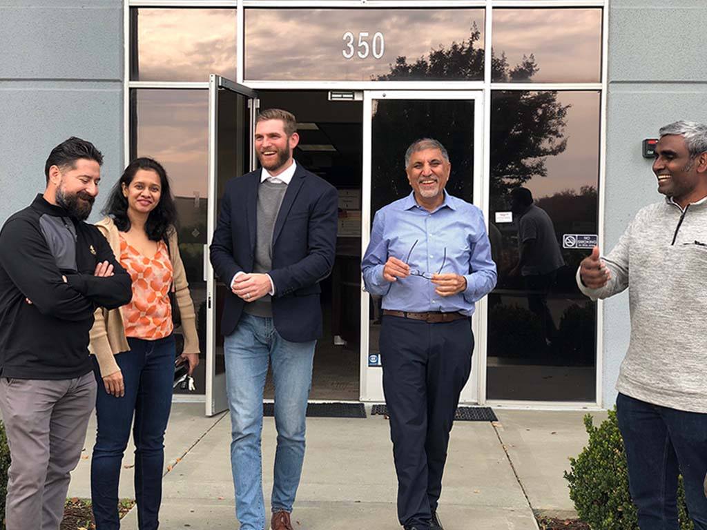Five team members from Acutrack stand smiling in front of their headquarters building numbered 350. The group includes a diverse mix of professionals - one wearing a black sweater, a woman in an orange patterned top with a beige cardigan, a tall man in a navy blazer and grey sweater, a man in a light blue button-down shirt, and another in a light-colored quarter-zip pullover.