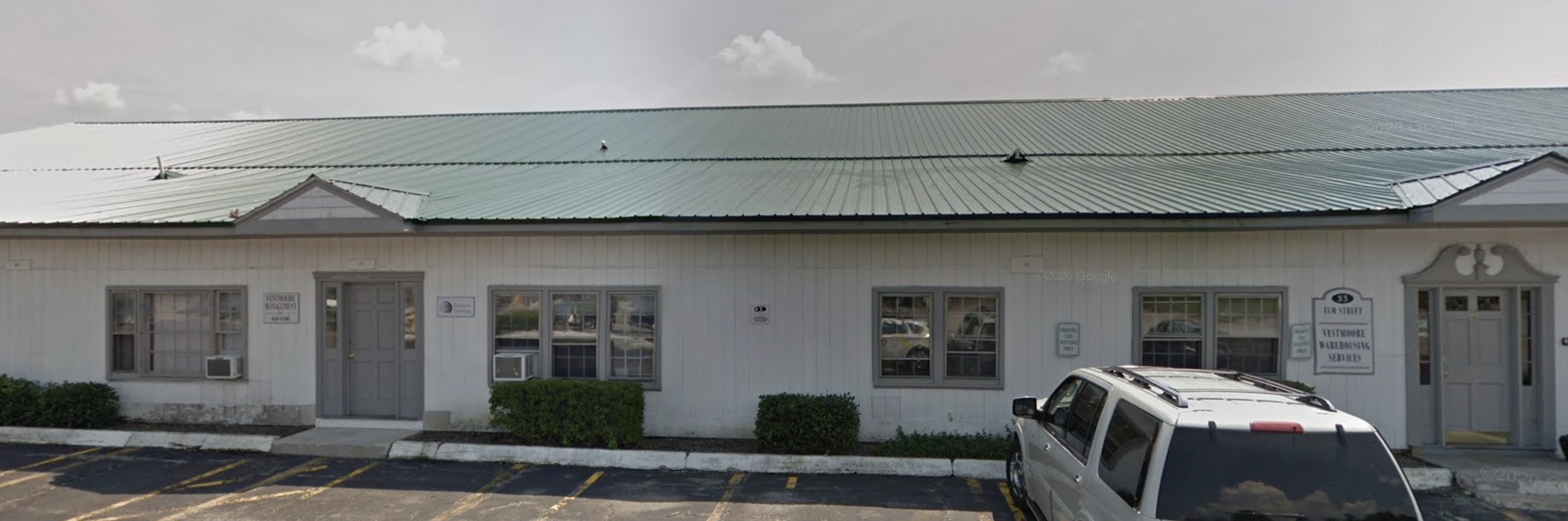 A single-story white building with a green metal roof. The structure has multiple windows and doors with grey trim, small shrubs planted along the foundation, and a parking area in front. The simple commercial building appears to be part of a business complex, with 'Law Street' visible on one of the office signs.