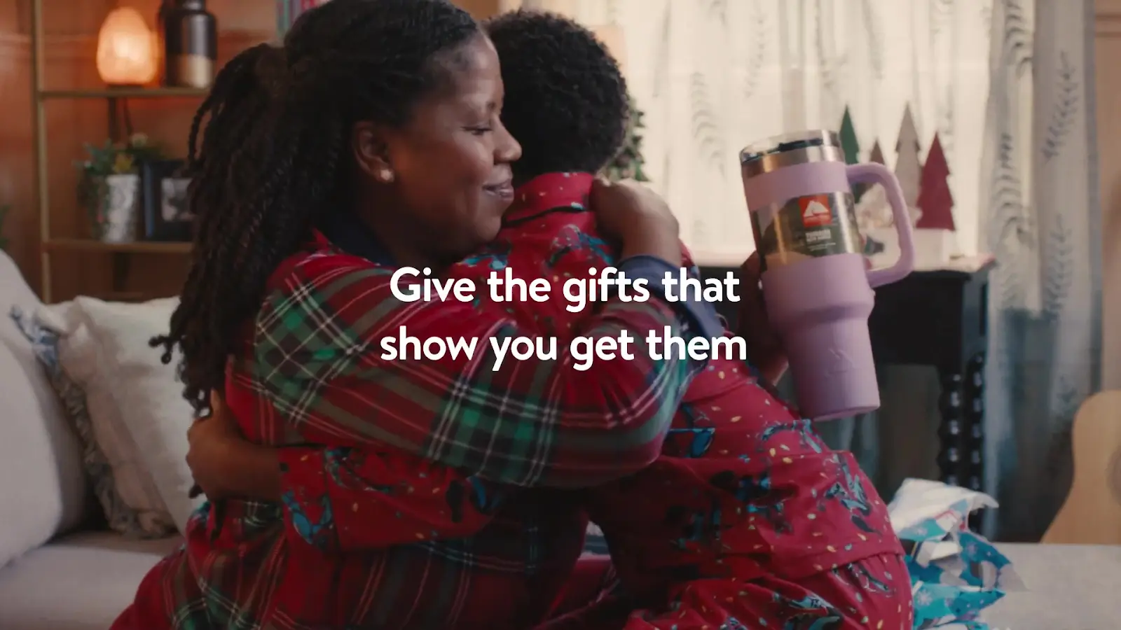 A joyful moment between a Black mother and child in a cozy living room setting, both dressed in festive pajamas and embracing, with a textual overlay saying "Give the gifts that show you get them." The scene suggests a warm, family-oriented holiday atmosphere.