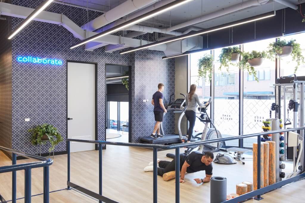 The open gym and rehabilitation floor at Upwell Health Collective, featuring a neon 'collaborate' sign, cardio machines, and clients exercising.