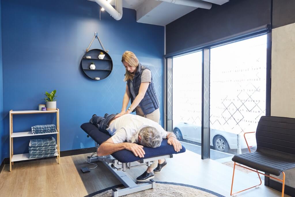 A physiotherapist performing a manual therapy treatment on a patient lying prone on a treatment table in a bright blue-walled clinical room at Upwell Health Collective.