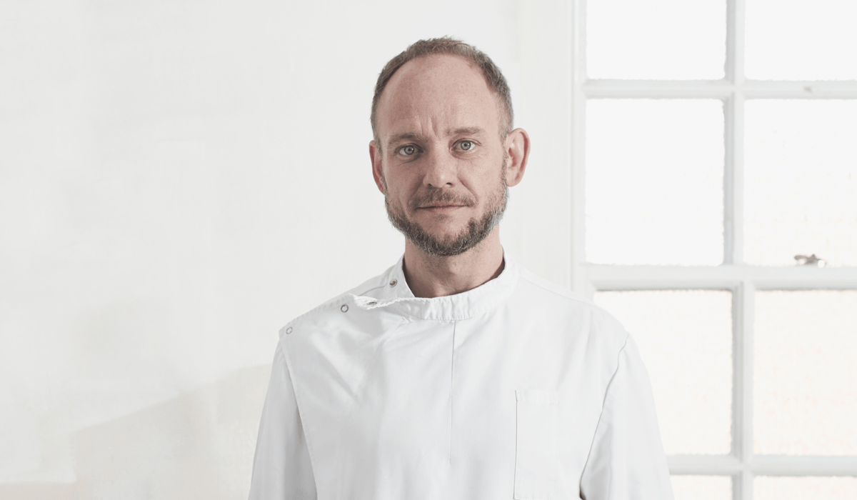 Portret van een man in een wit ziekenhuishemd, staande tegen een lichte achtergrond; hij is geïdentificeerd als Danny Orchard, eigenaar van Core Clapton Osteopathy.