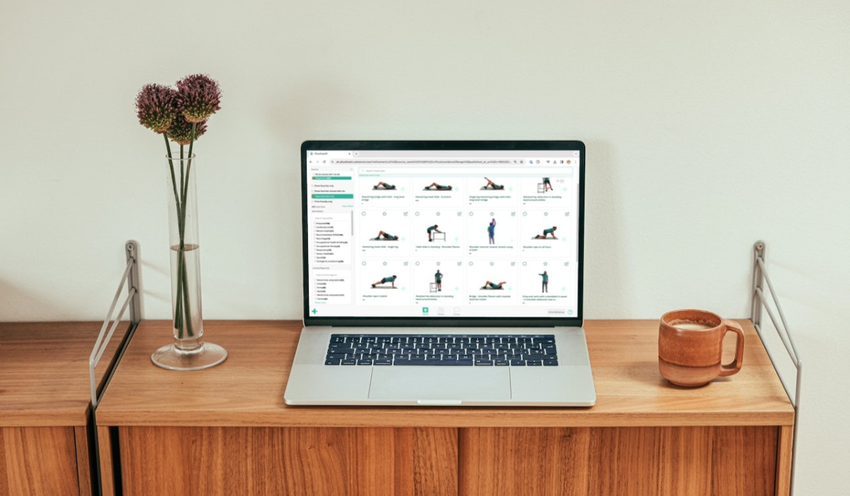 Sebuah laptop di atas meja kayu yang menampilkan antarmuka web perpustakaan Physitrack dengan kisi-kisi ilustrasi latihan.