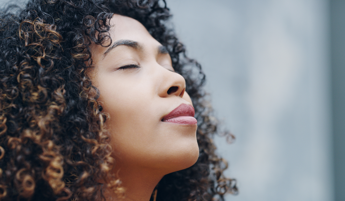 Close-up portrait of a woman with curly hair and closed eyes appearing calm and meditative outdoors.