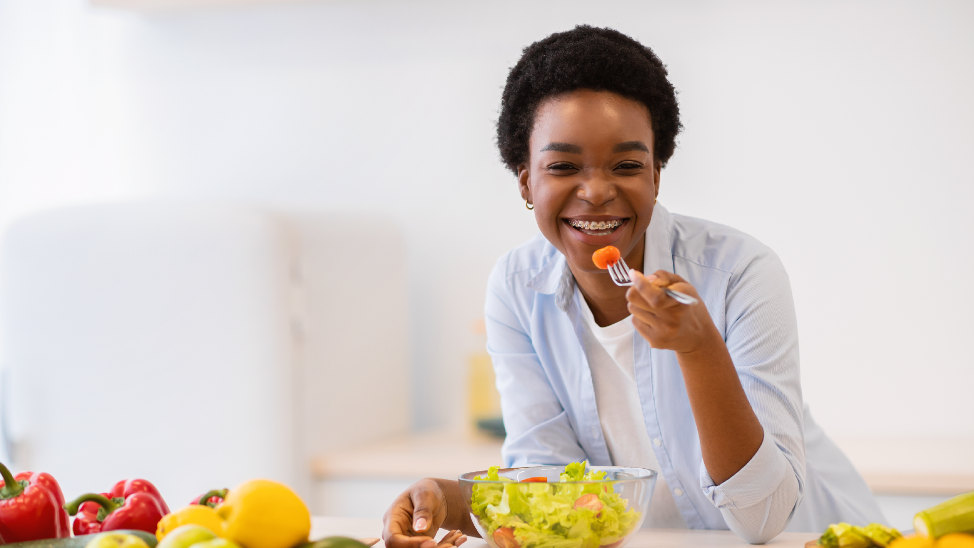 Smiling woman eating a fresh salad with colourful vegetables on the counter in a kitchen.