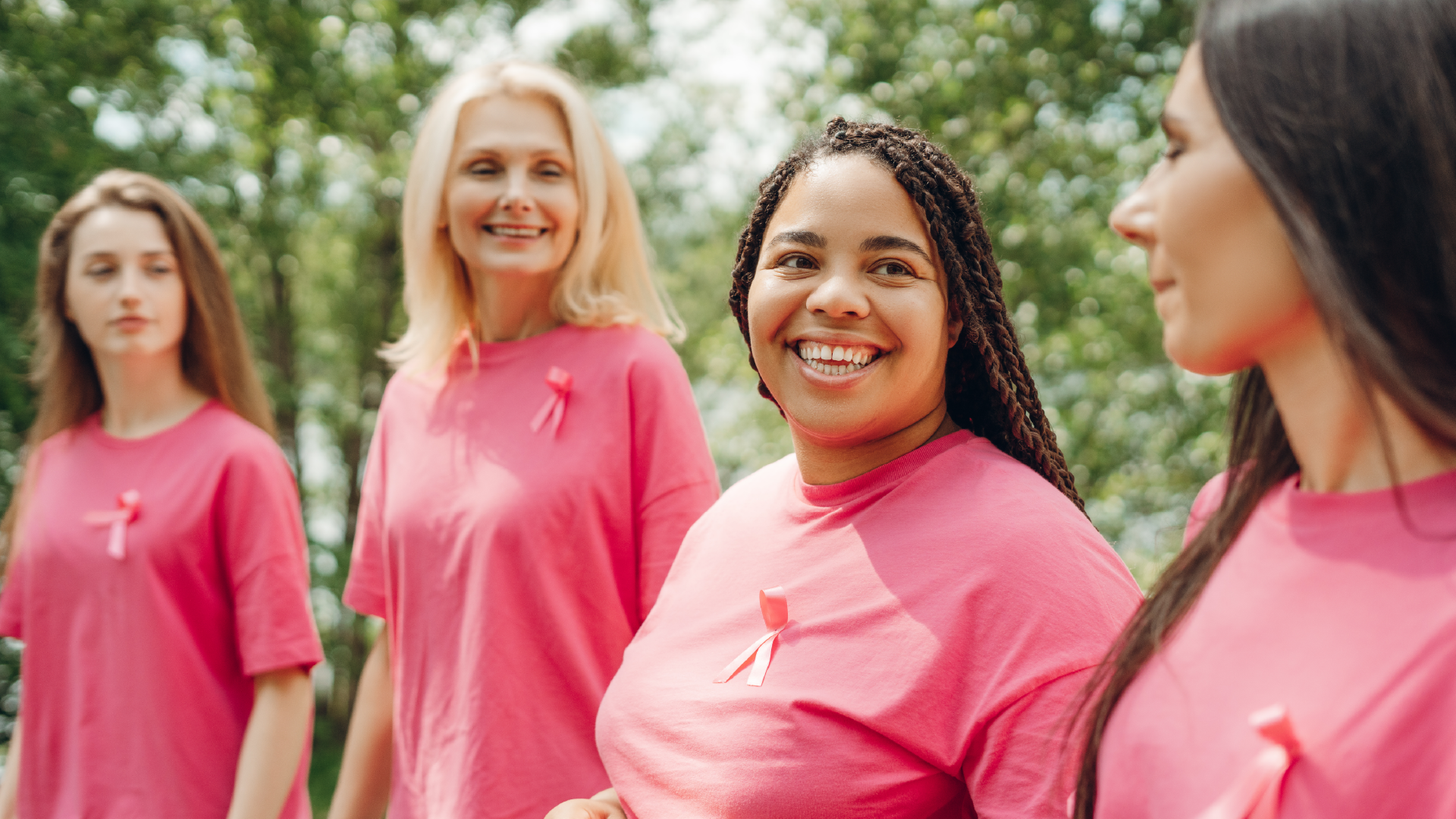 Four smiling women in pink t-shirts with breast cancer awareness ribbons standing outdoors