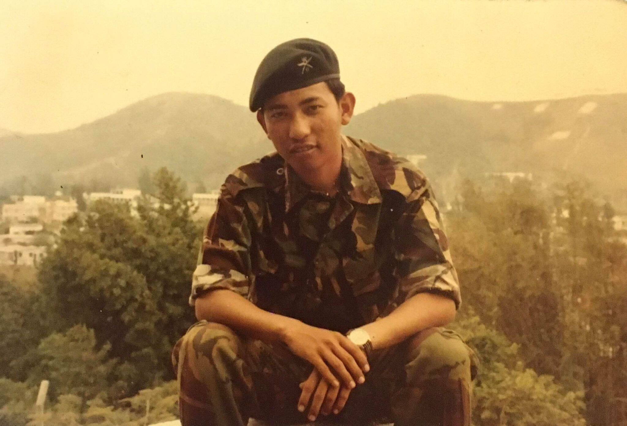 Vintage photo of a young man in camouflage uniform and beret seated before hills and trees