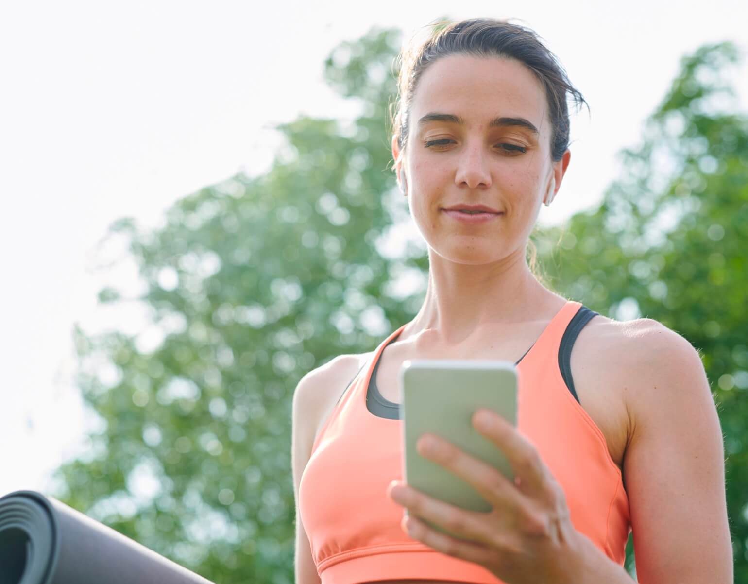Woman in an orange tank top with earbuds and a yoga mat looking at her phone outdoors