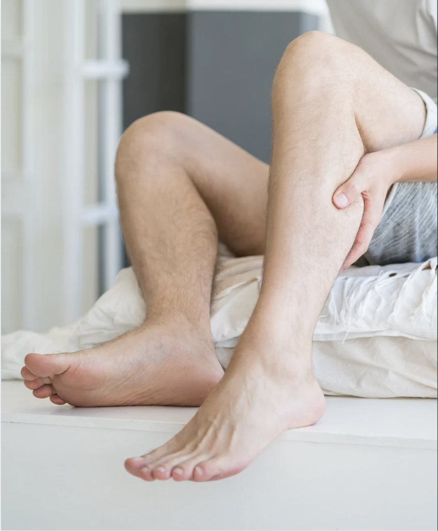 Person seated on a bed gripping their lower leg and ankle, suggesting foot or ankle pain