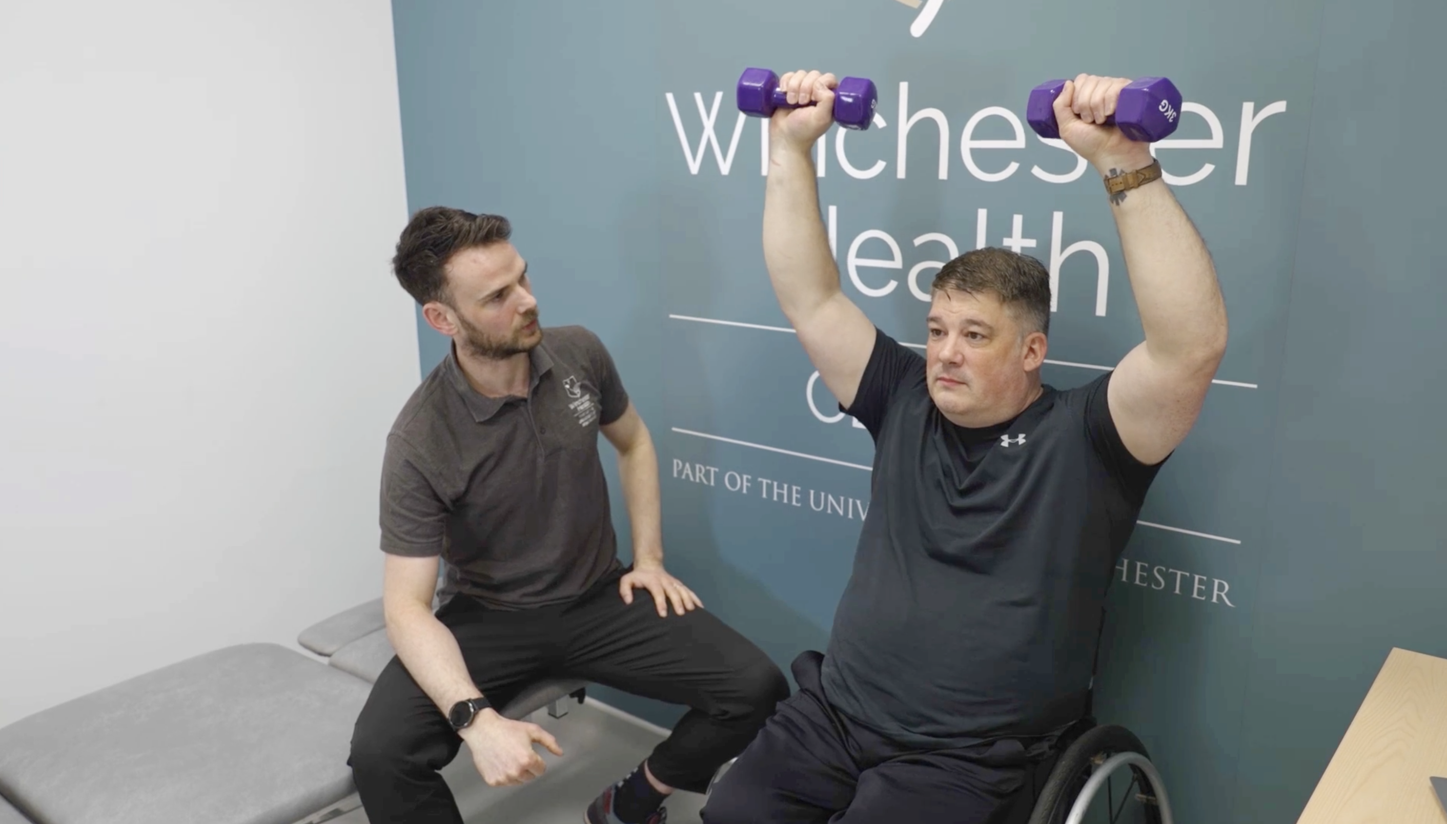 Man in a wheelchair lifting purple dumbbells overhead as a Winchester Health clinician watches.
