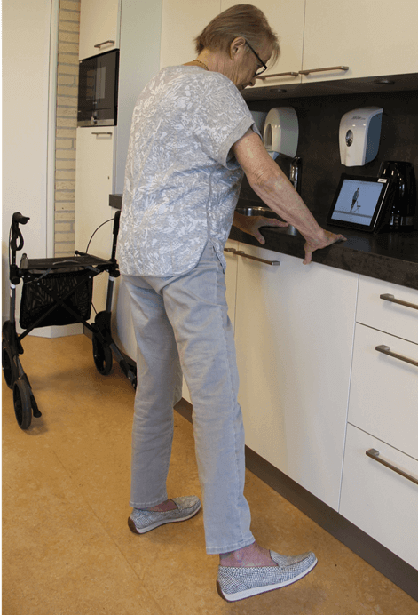 Eine ältere Frau, die mithilfe eines an der Wand befestigten Tablets eine Übung mitmacht, während sie in einer Küche steht und neben ihr ein Rollator steht