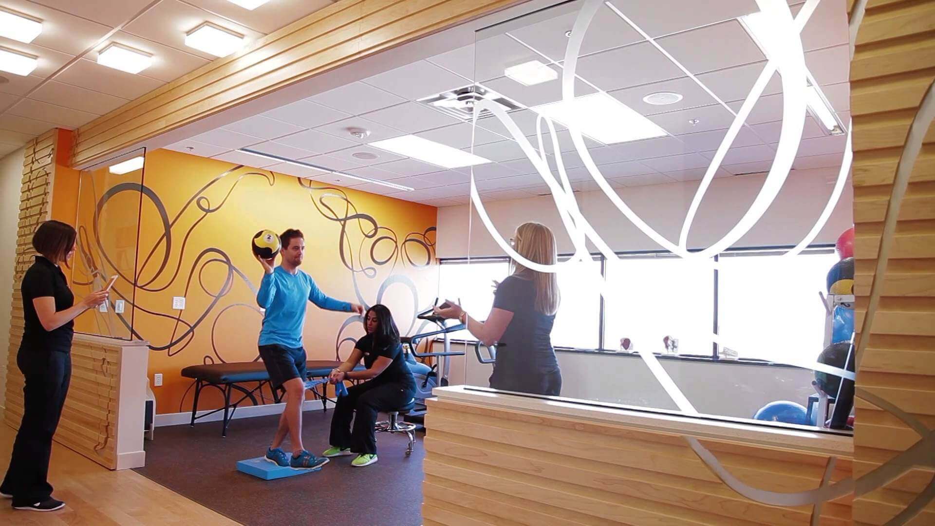 Bright physiotherapy gym with patients and therapists training near a swirl-patterned glass partition.