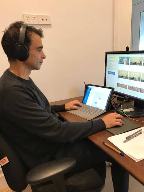 Man wearing headphones working at a desk with a laptop and monitor showing exercise videos