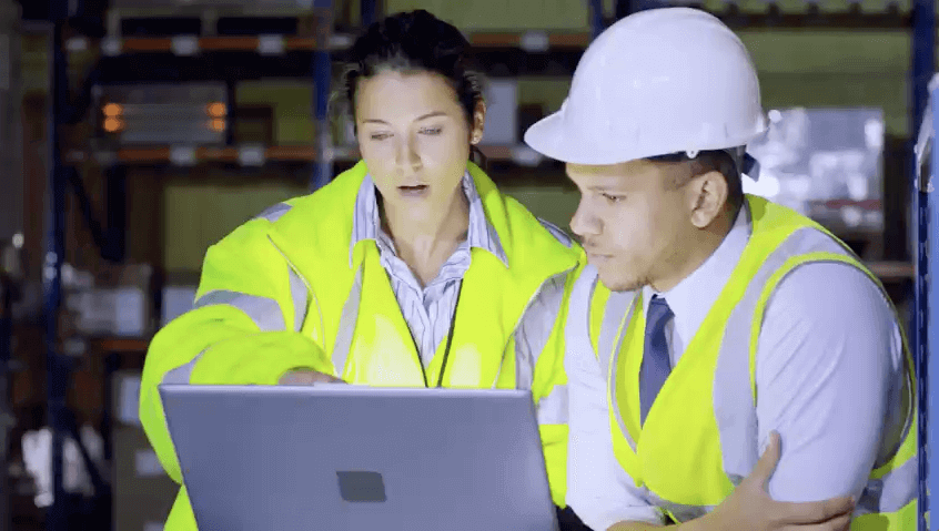 Man and woman in hi-vis safety vests looking at a laptop together in a warehouse.