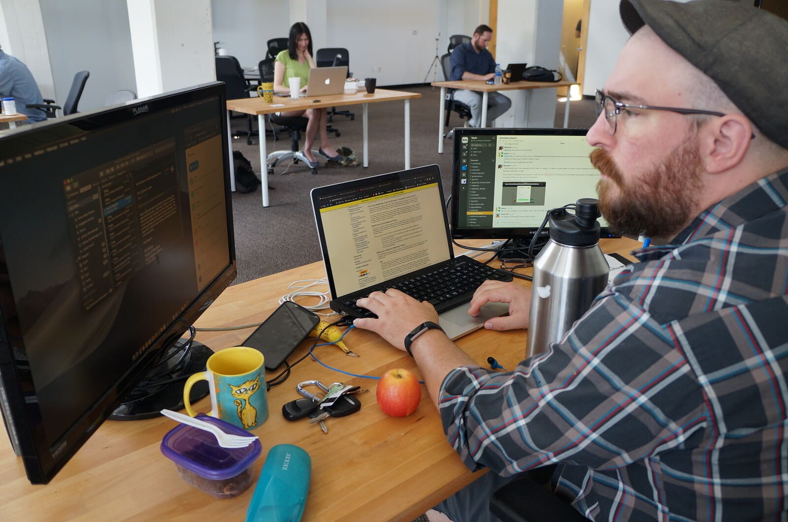 Developer coordinating with a distributed team over Slack across a laptop and two monitors