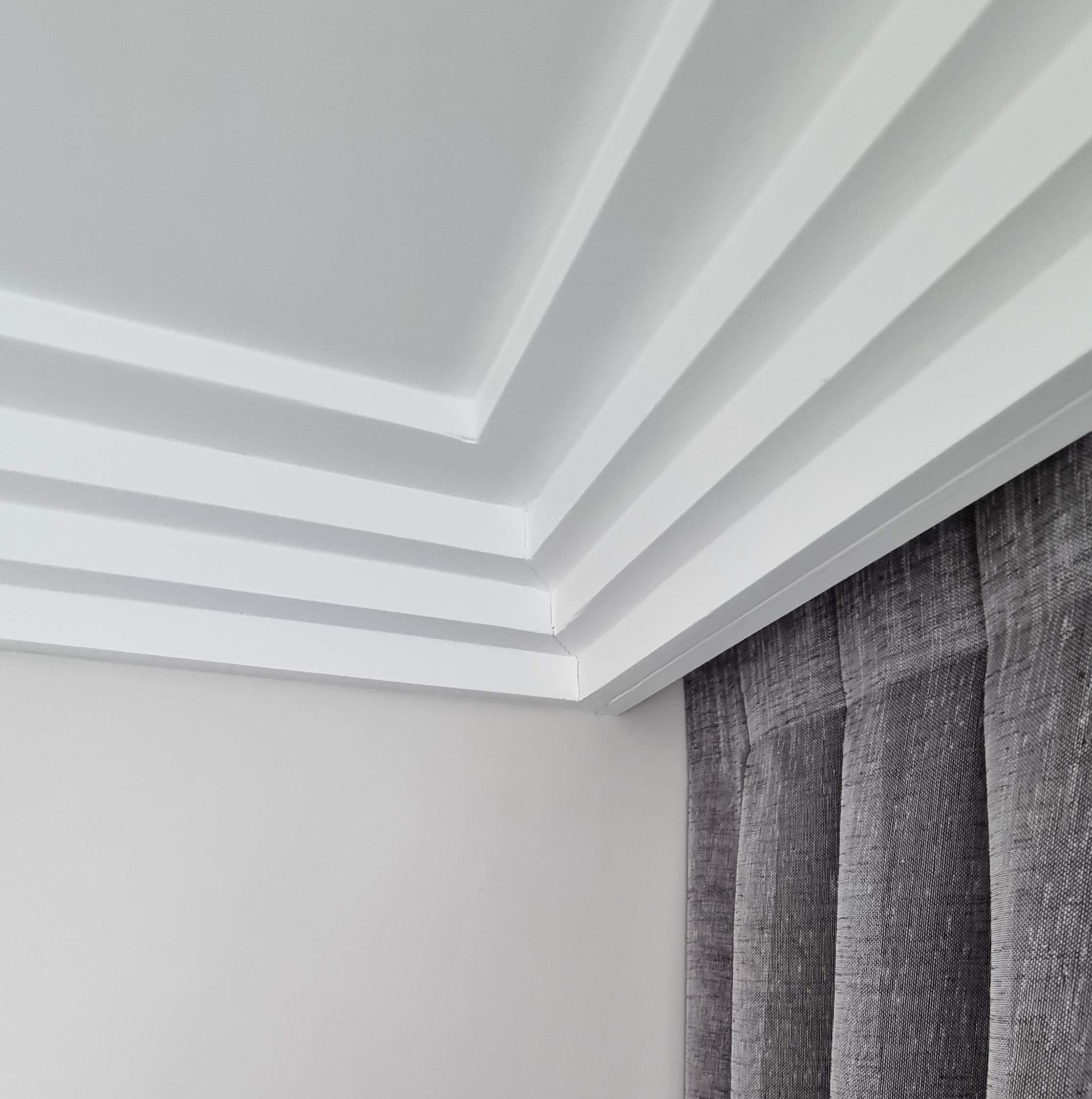 Corner of a room showing multi-layered white ceiling molding and gray textured curtains.