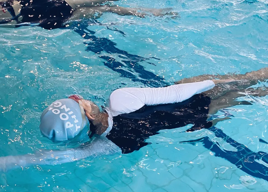 戴印有 POOL THINGS 字樣淺藍泳帽的學員在泳池練習背泳，旁有其他泳者。