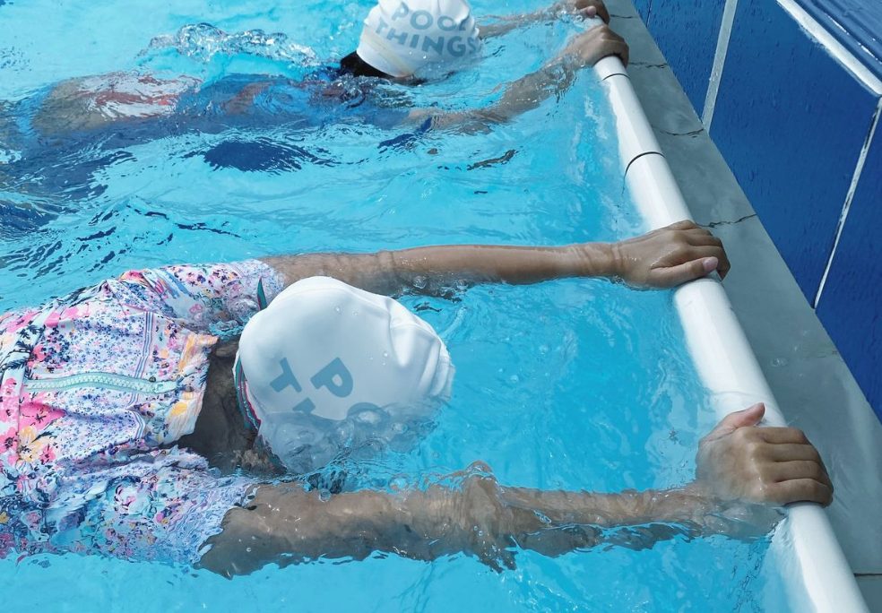 兩位戴白色 POOL THINGS 泳帽的學員趴在池邊扶欄，練習伸展與浮水姿勢。