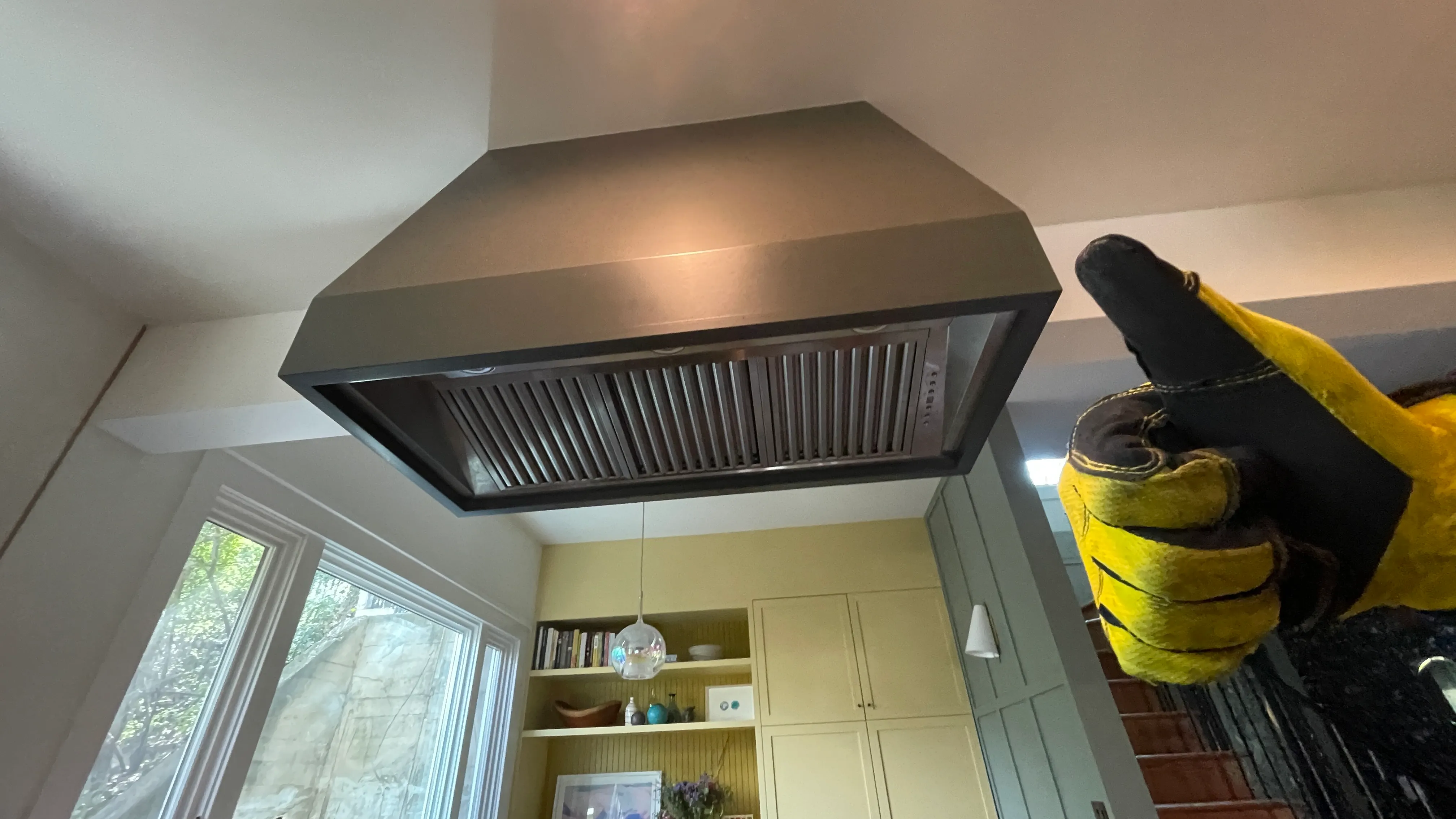 Close-up of a stainless steel kitchen vent hood installed on a ceiling with yellow cabinets and windows in the background, a hand wearing a yellow and black glove giving a thumbs up to the right.
