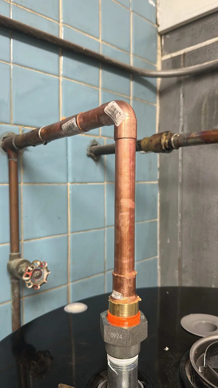 Close-up of copper plumbing pipes with a brass fitting and valve against a blue tiled wall.