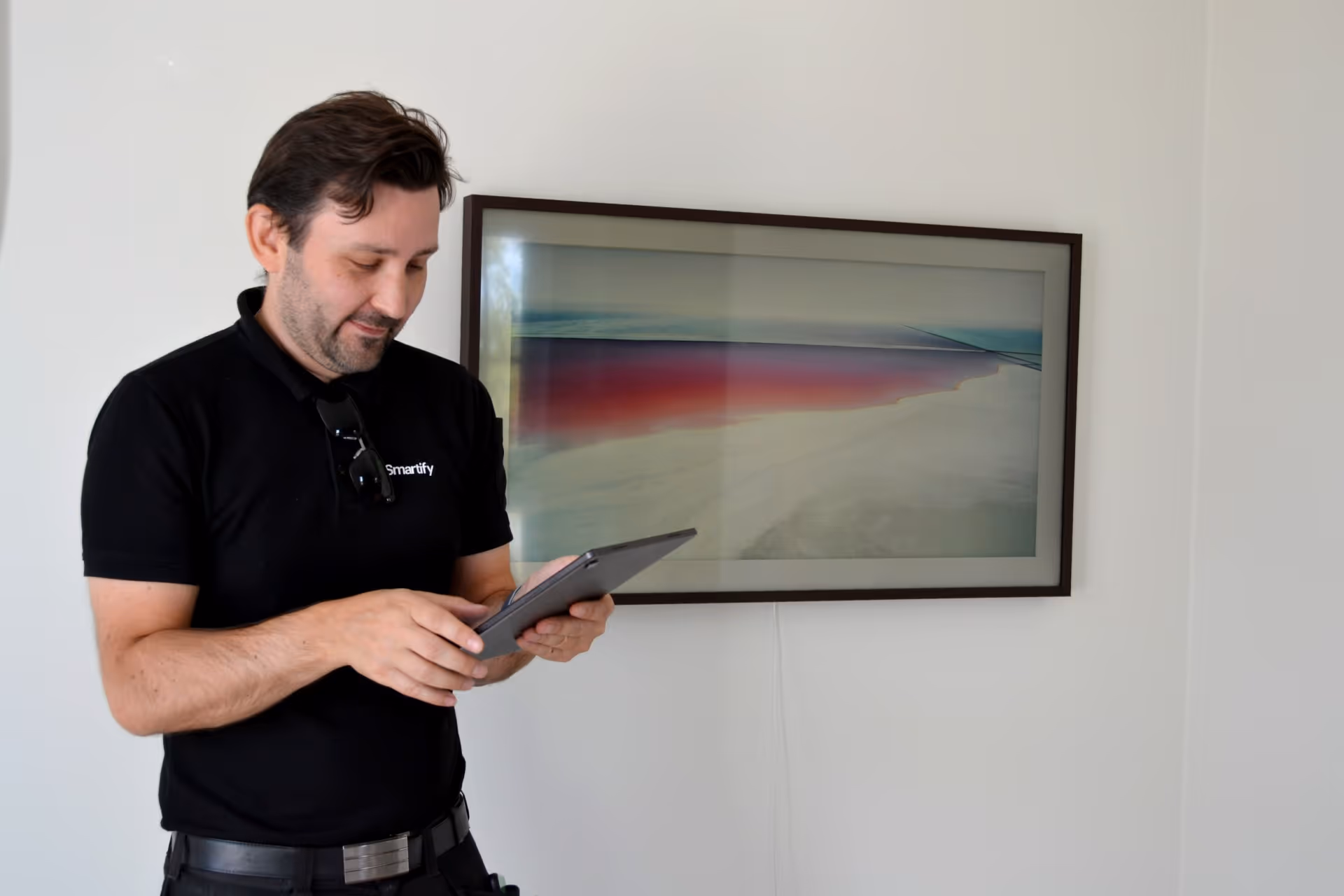 Man in black Smartify shirt using a tablet in front of a landscape painting on a white wall.