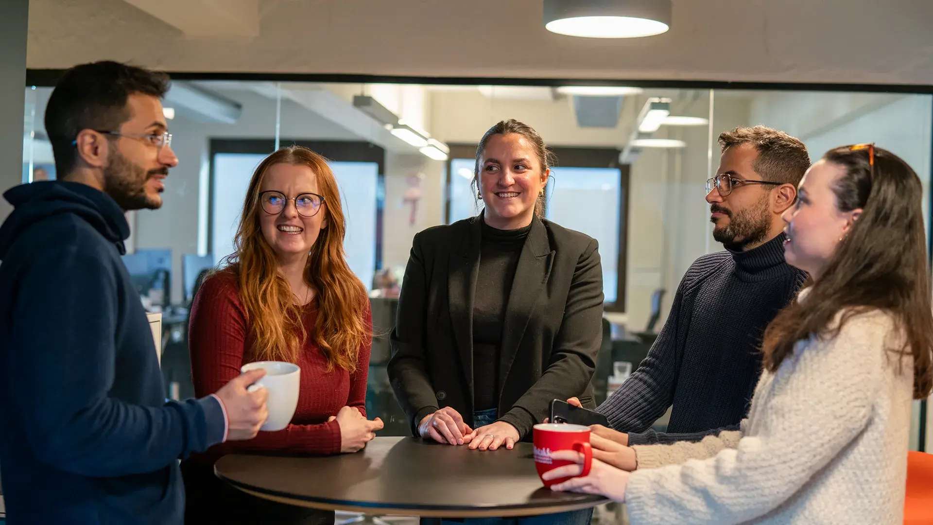 Fem personer som har kaffeprat rundt et bord på kontoret