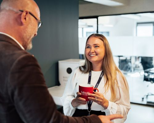 Kvinne med rød kaffekopp smiler til mann som står med ryggen til