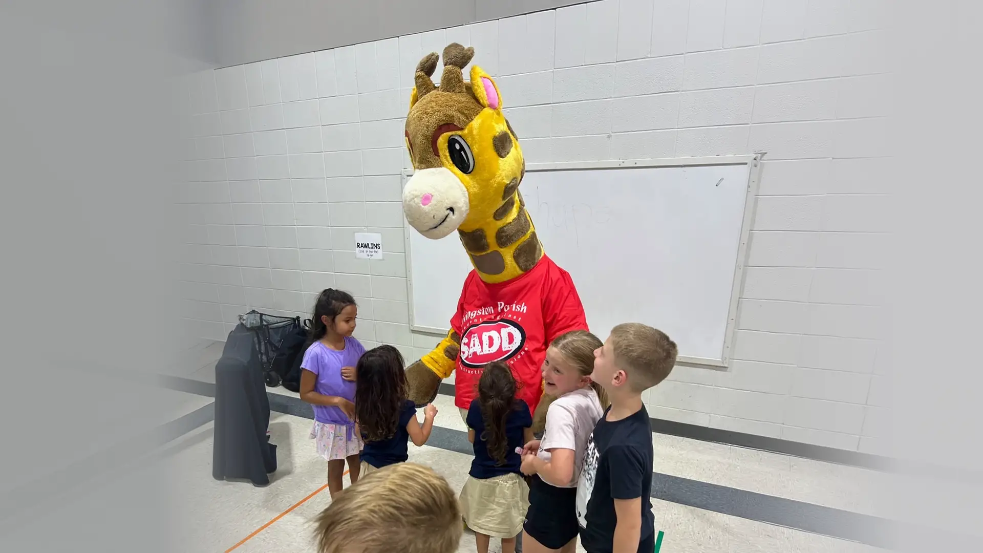 Children gathered around a person in a giraffe mascot costume wearing a red shirt with 'SADD' on it inside a room with tiled floors and a whiteboard on the wall.