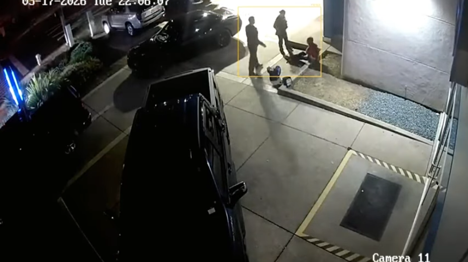 Nighttime CCTV view of three people near a building entrance, with one person sitting on the ground and two standing nearby.