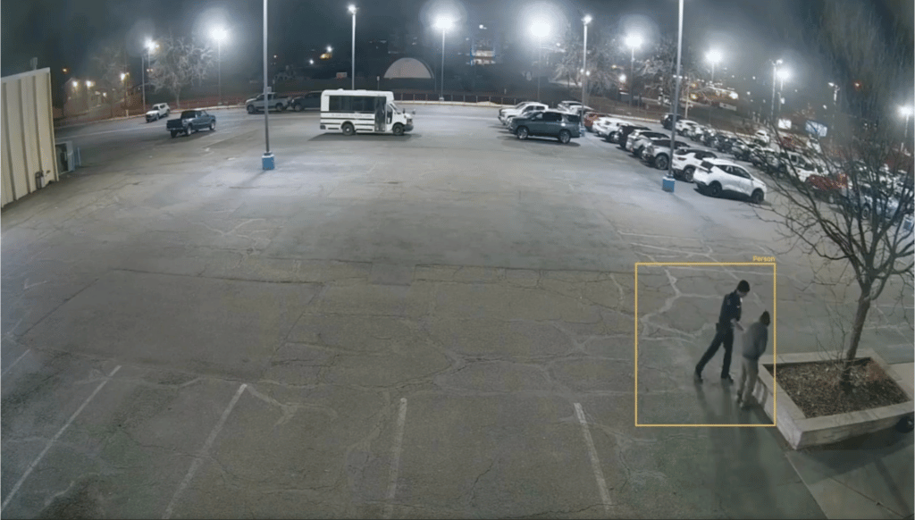 Night view of a mostly empty parking lot with a white shuttle bus and two people near a tree on the right side.