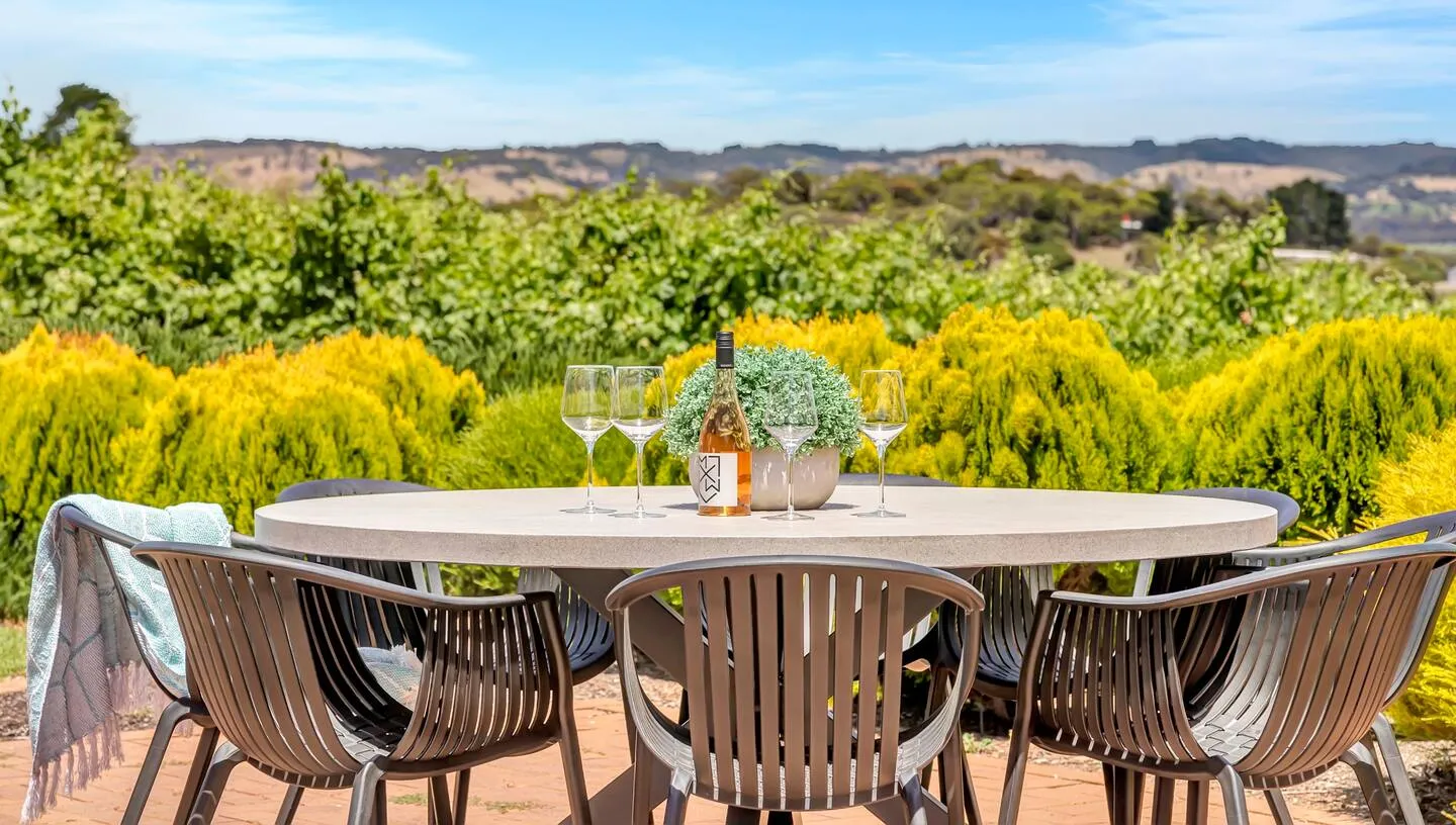 An outdoor table surrounded by chairs, with four wine glasses and a bottle of pink wine on top. The property is at Golden Homestead, an Airbnb in McLaren Vale, South Australia