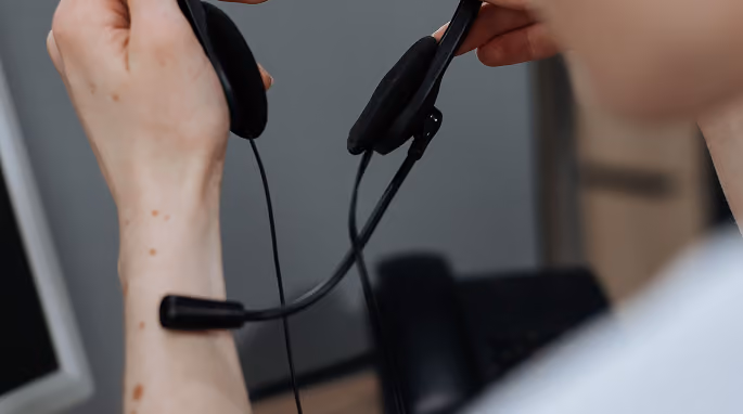 Person holding a black headset with a microphone near a computer monitor and phone.