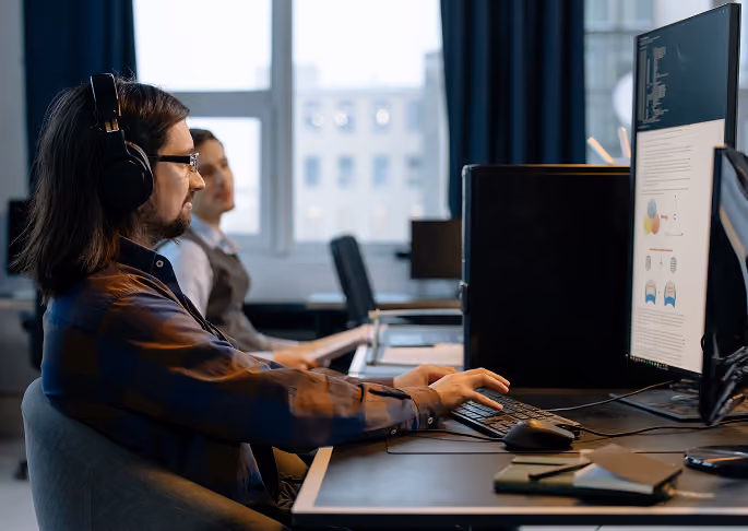 Two men working on computers in an office, one wearing headphones and glasses.