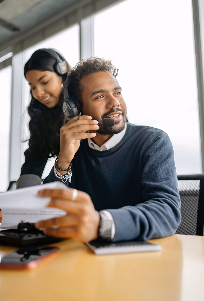 Man wearing headset and holding papers sits at desk while woman wearing headset stands behind him smiling.