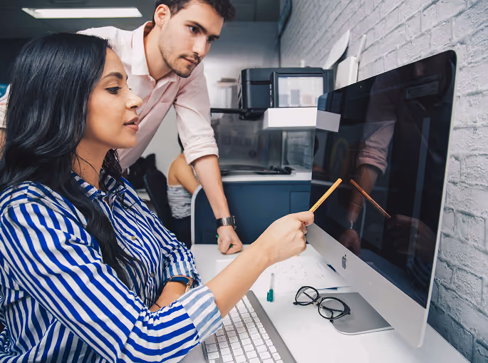 Woman pointing at a computer screen with a pencil while a man watches attentively in an office.