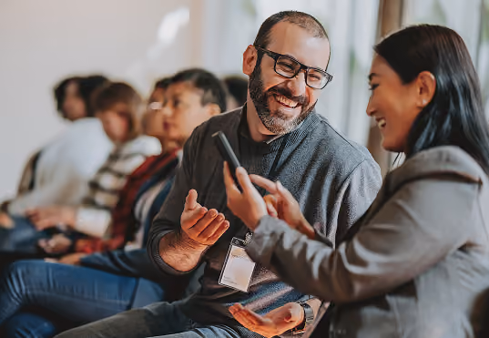 Smiling man wearing glasses talks with a woman holding a smartphone during a group meeting.