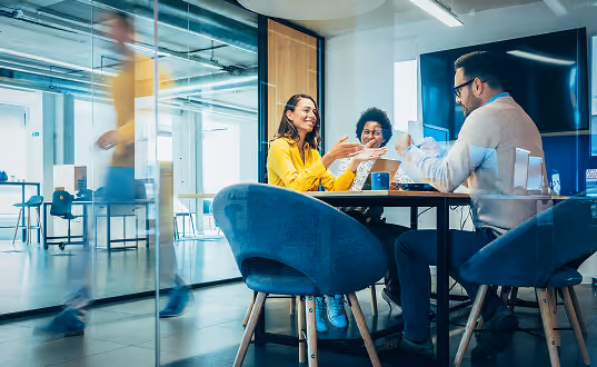 Three colleagues smiling and gesturing during a casual meeting in a modern glass-walled office.