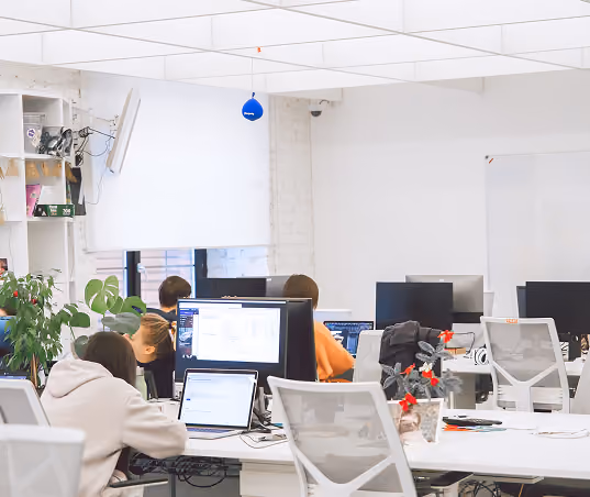 Modern office with people working at desks using laptops and monitors under white ceiling panels.